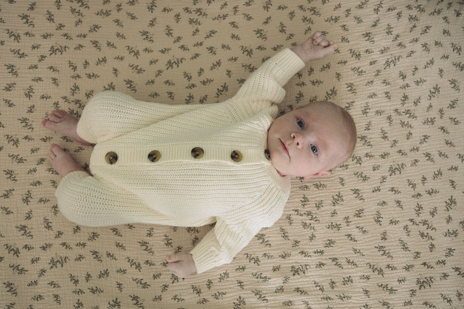 A baby wearing a cream-colored sweater lies on a floral patterned surface in different poses.