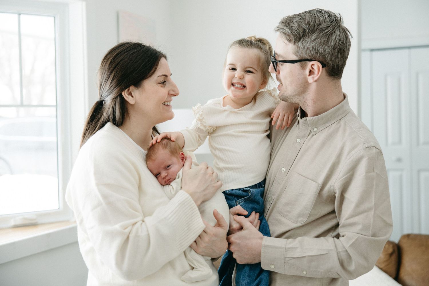 A joyful family wearing cream colored sweaters shares tender moments together in a bright white room.