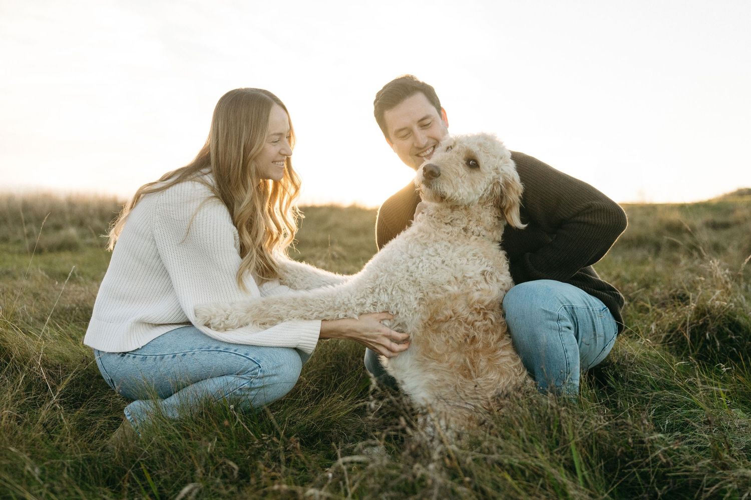 A couple kneels on a grassy hillside at sunset, smiling as they pet a fluffy golden doodle dog.