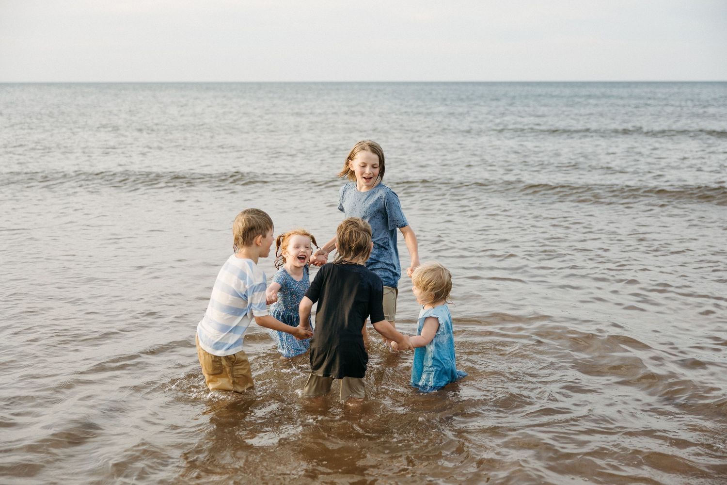 Five young children laughing and playing together in shallow ocean water on a beach.