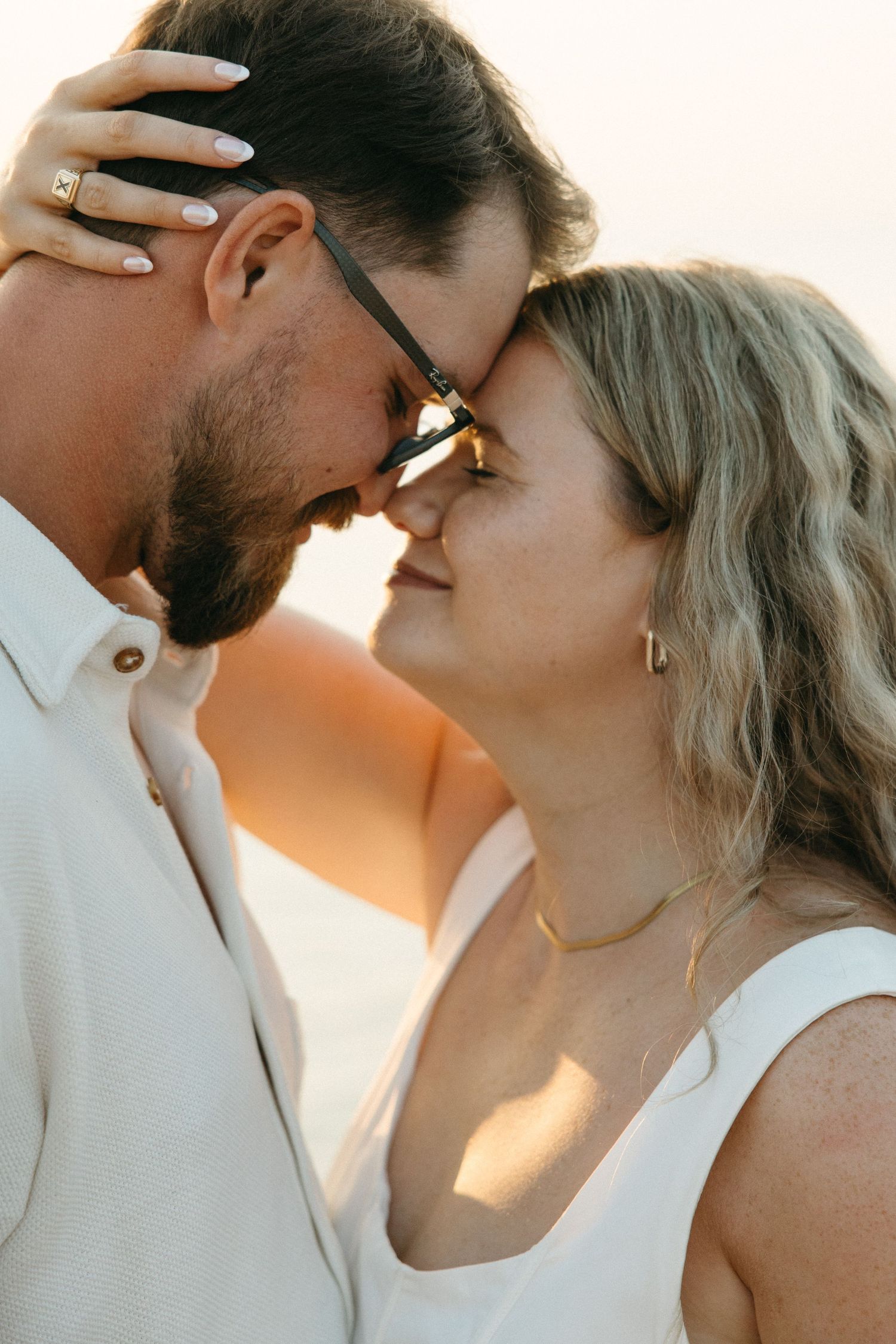 Romantic couple touching foreheads at golden hour sunset, woman wearing sunglasses and gold necklace.