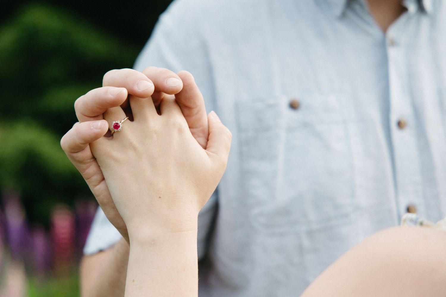 Close-up of a couple holding hands outdoors, woman wearing a red gemstone ring, with purple flowers in background.