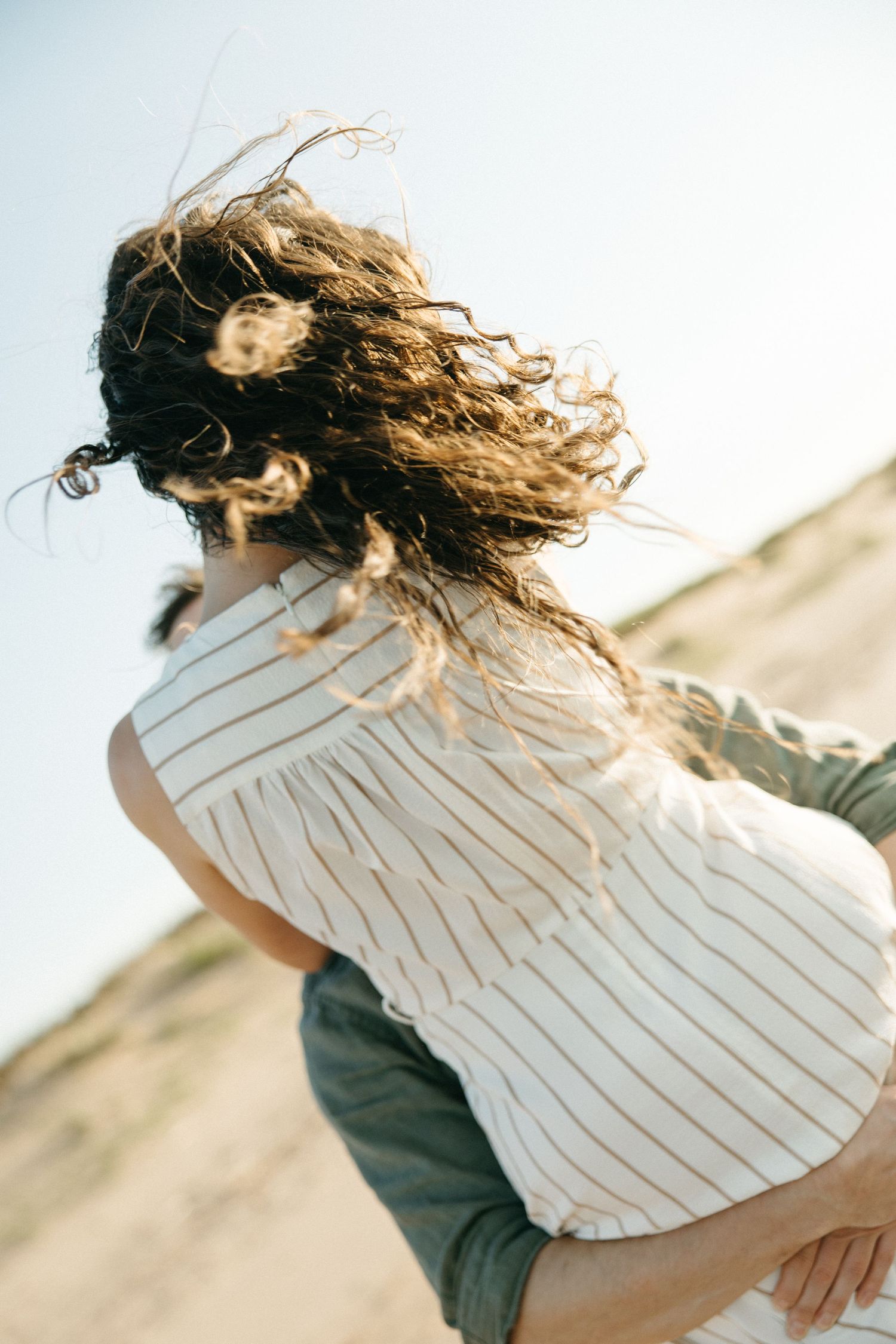 Man lifting woman in striped dress on sunny beach, her curly hair blowing in wind.