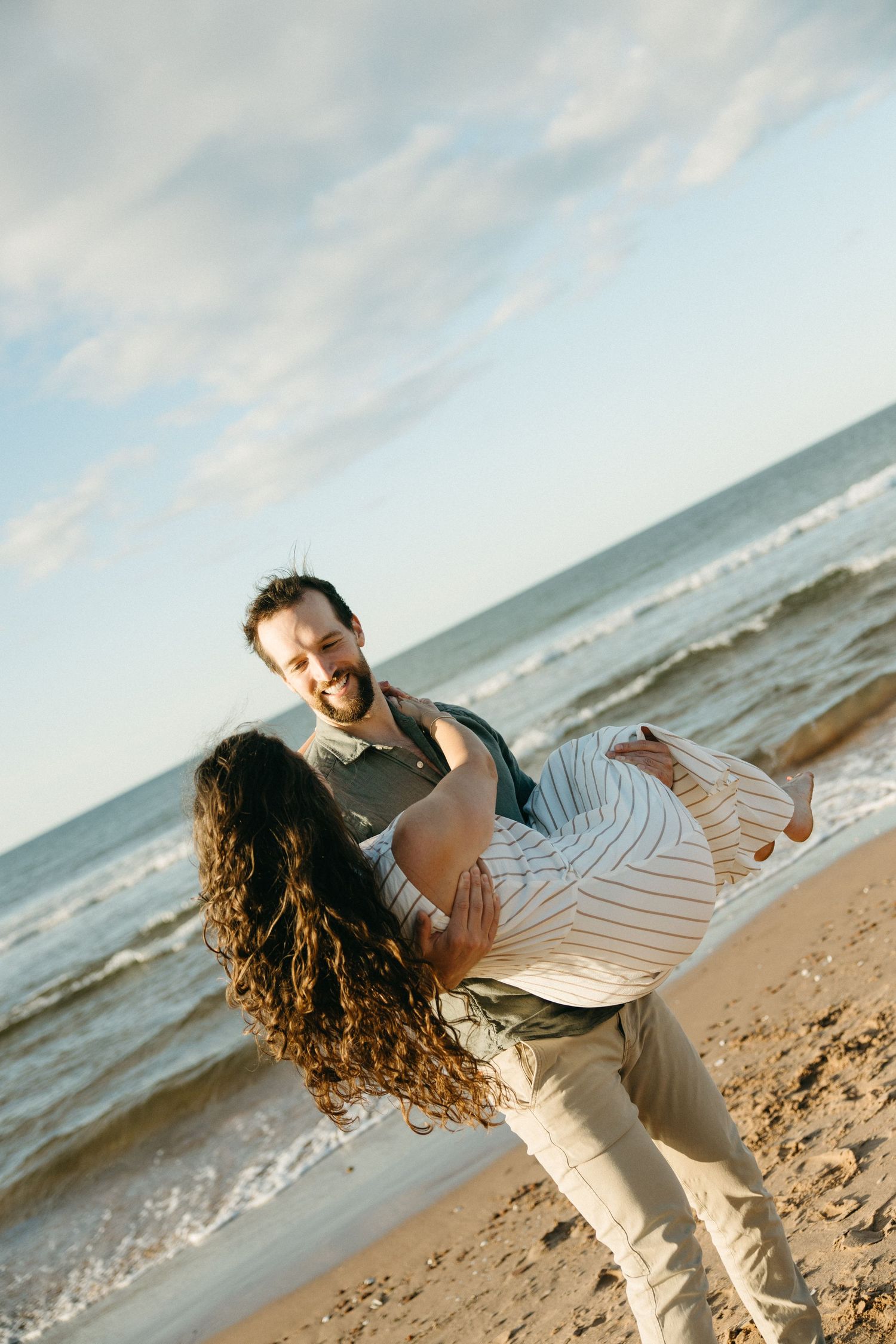 Smiling bearded man lifting woman with curly hair on sandy beach with ocean waves behind them.
