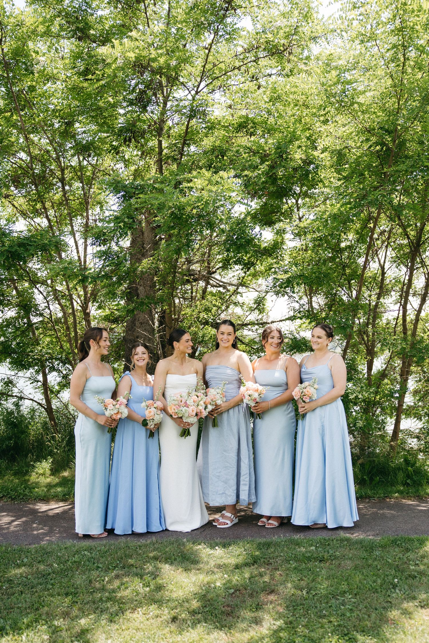 Bride in white gown with five bridesmaids in light blue dresses holding floral bouquets outdoors among green trees.