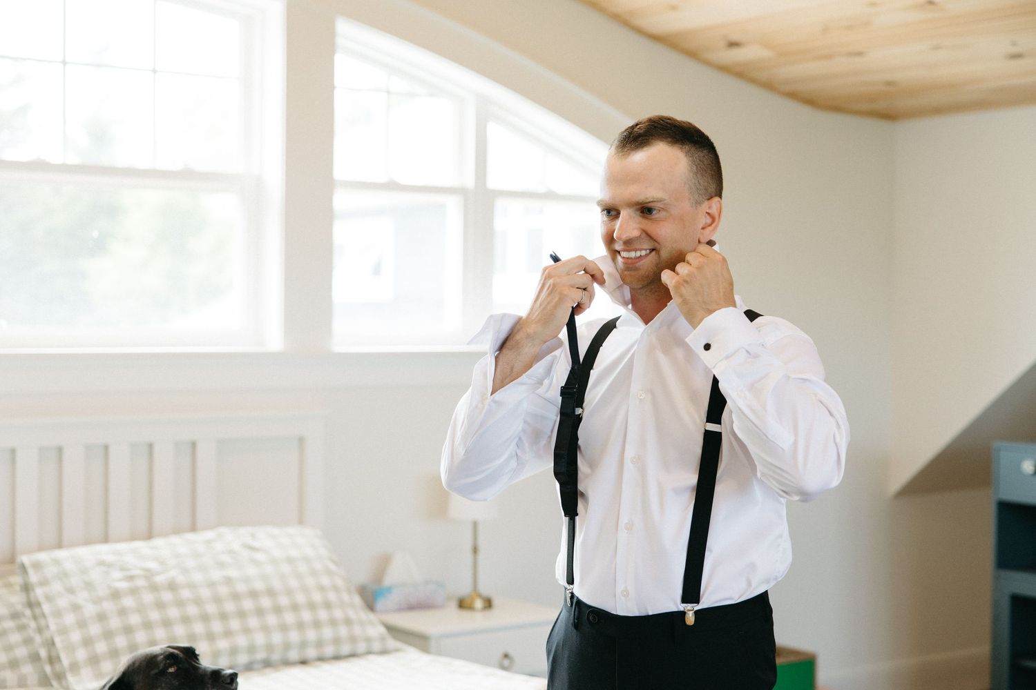Groom in white shirt and suspenders adjusting his bow tie while getting ready in a bright bedroom.