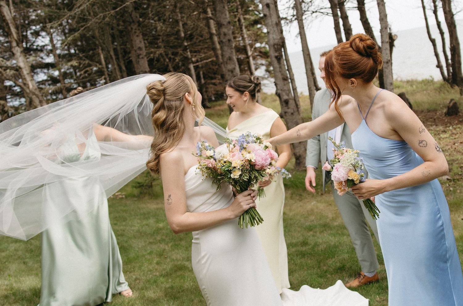 Bride in white gown holding colorful bouquet with bridesmaids in blue dresses adjusting her veil outdoors.