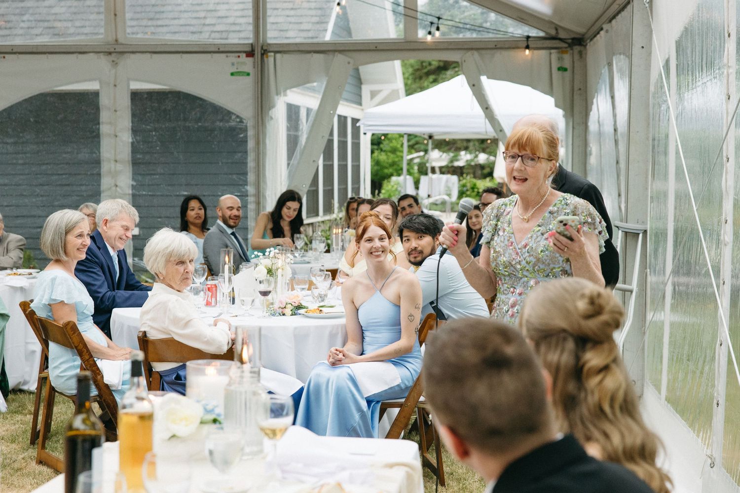 Woman in floral dress gives a speech at an elegant tented wedding reception with guests seated at decorated tables.