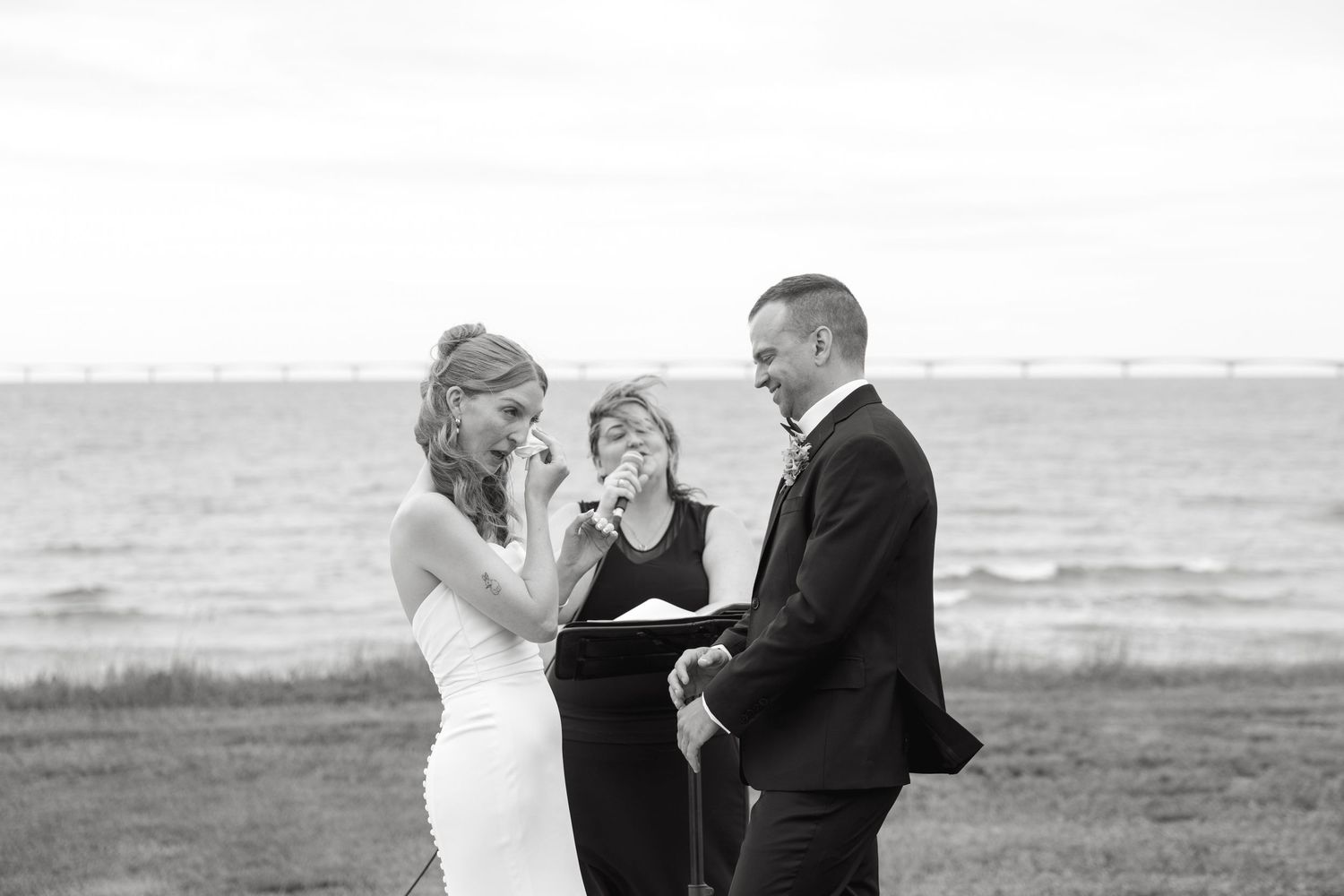 Bride wipes tears during emotional outdoor wedding ceremony by the water with officiant behind.