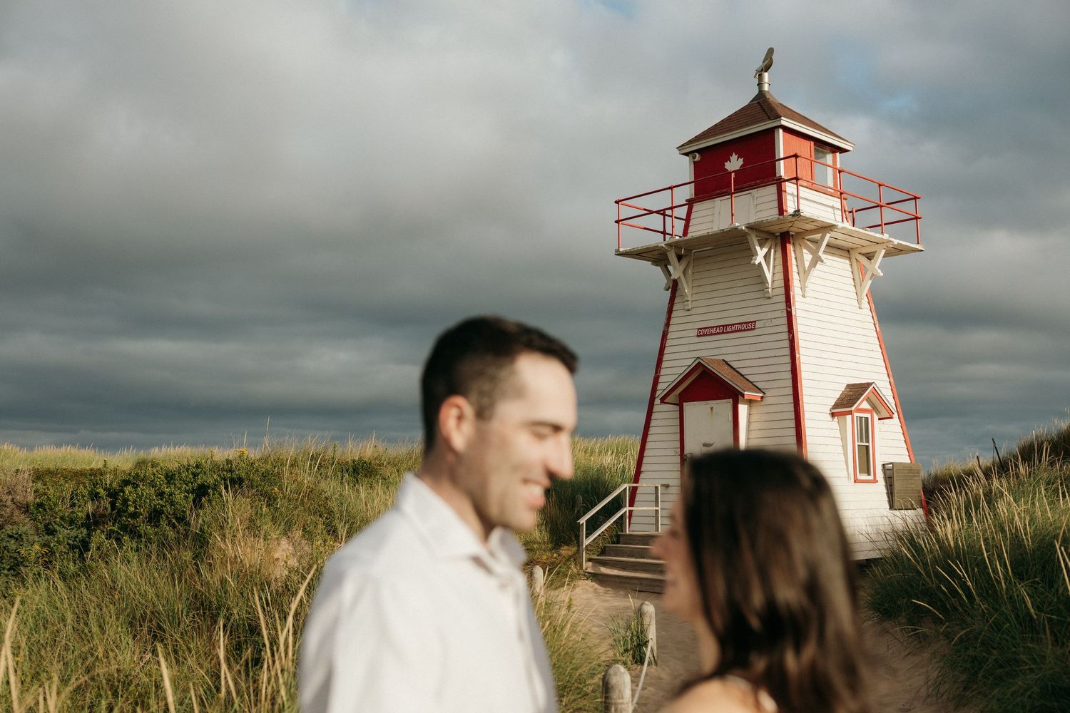 Wedding couple walks near coastal lighthouse under dramatic cloudy sky.