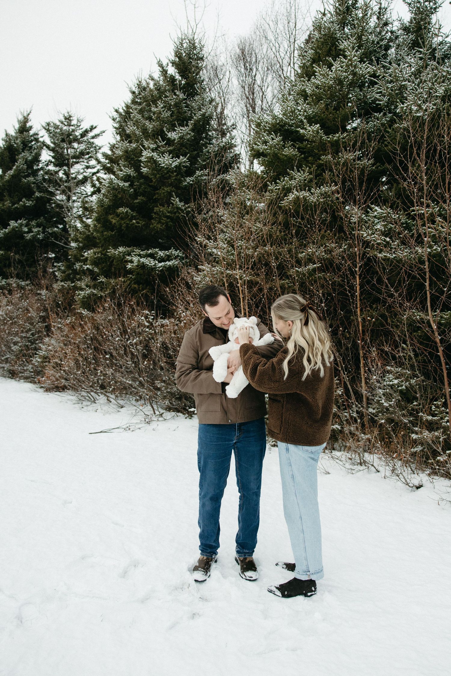 A couple in matching brown coats holds their baby while standing in fresh snow surrounded by evergreens.