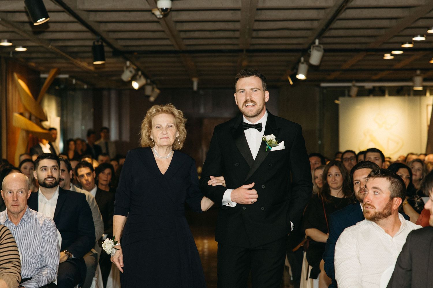Wedding processional in an indoor venue with guests seated on white chairs.