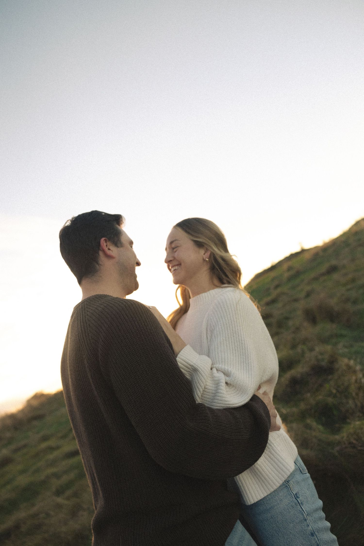 Smiling couple embracing on a grassy hillside at sunset, wearing cozy sweaters in a warm, romantic outdoor moment.