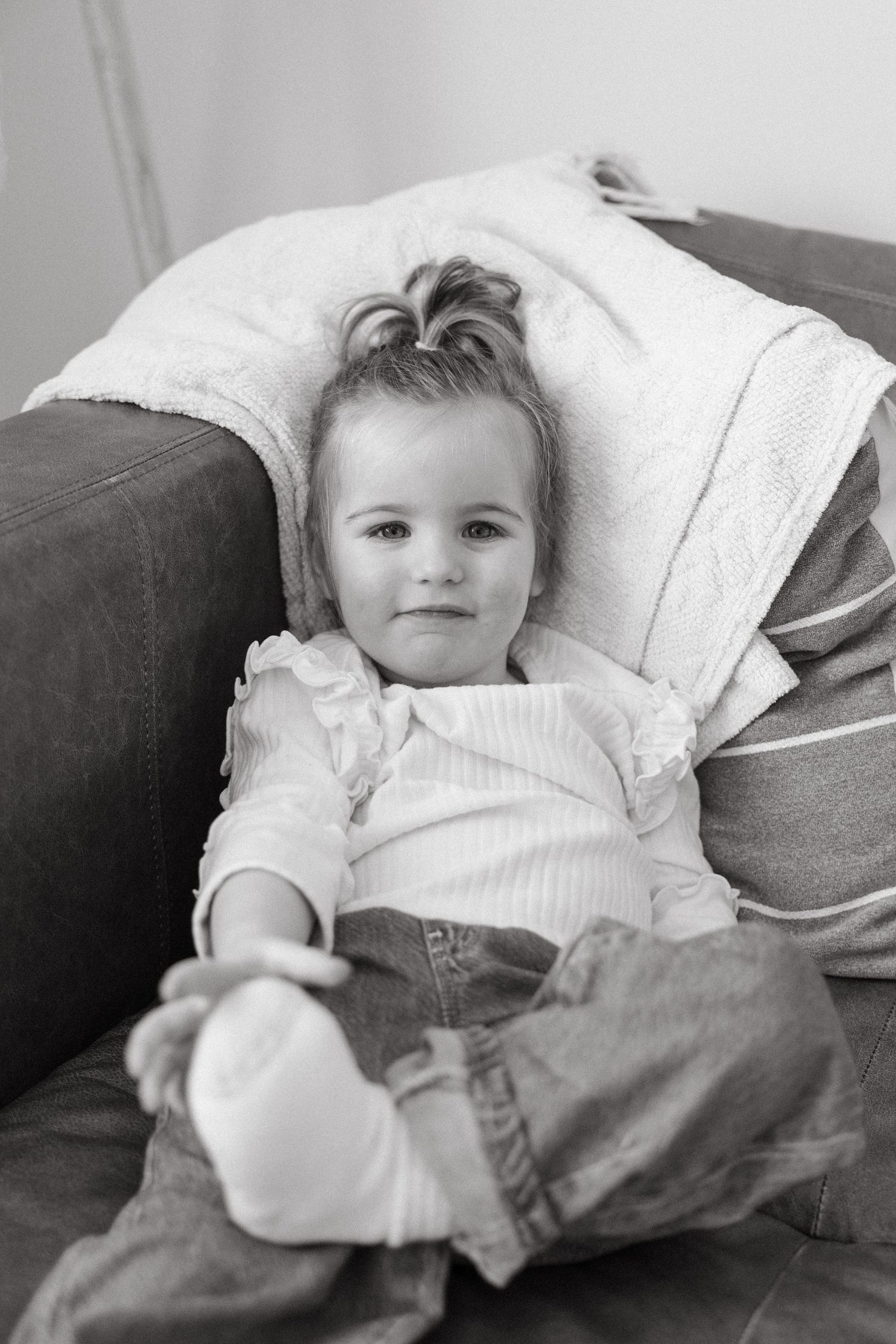 A child rests comfortably on a grey couch wrapped in blankets with a sweet expression.