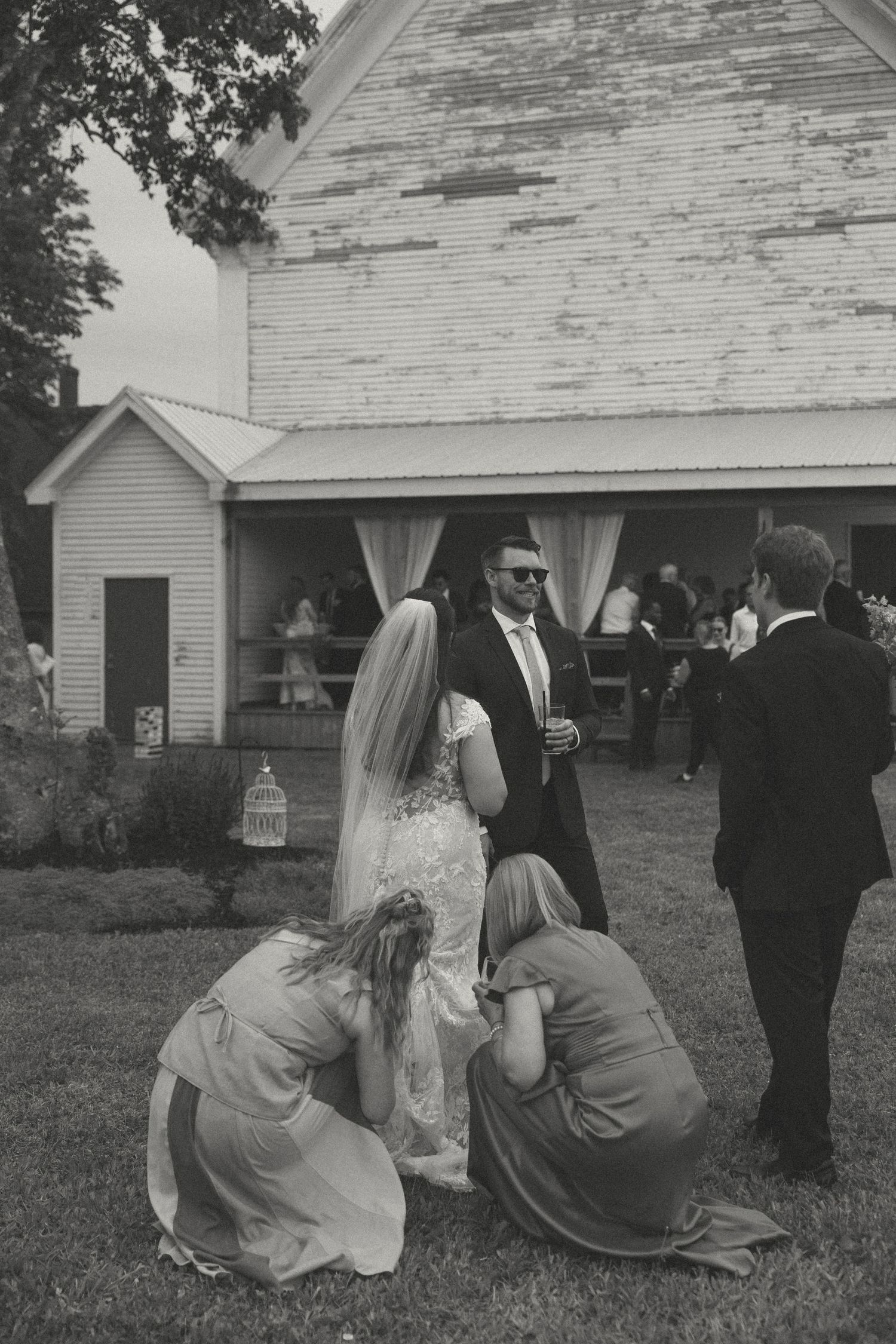 Wedding party members sitting on grass in front of a white house during outdoor festivities.