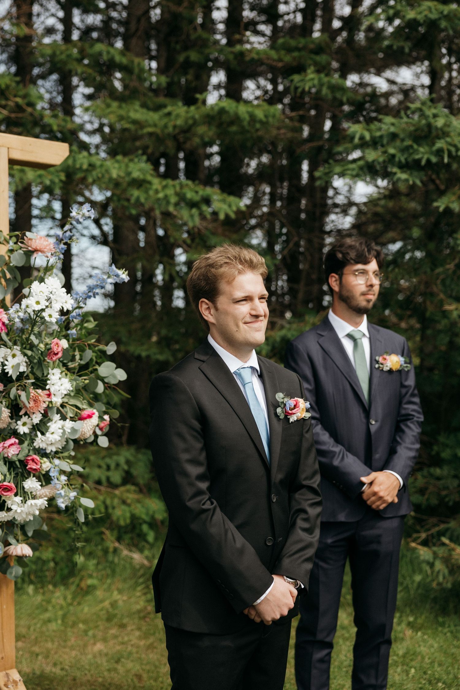 Groomsmen stand at decorated floral arch during outdoor wedding ceremony.
