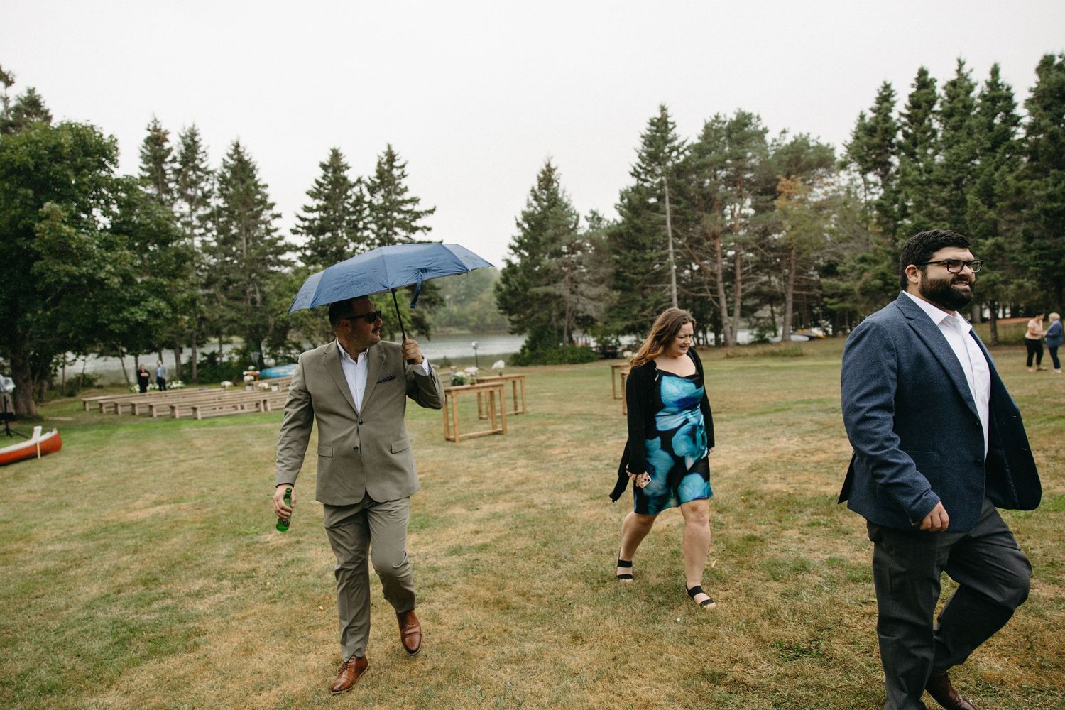 Wedding guests walk across a grassy field with one holding a blue umbrella on an overcast day.