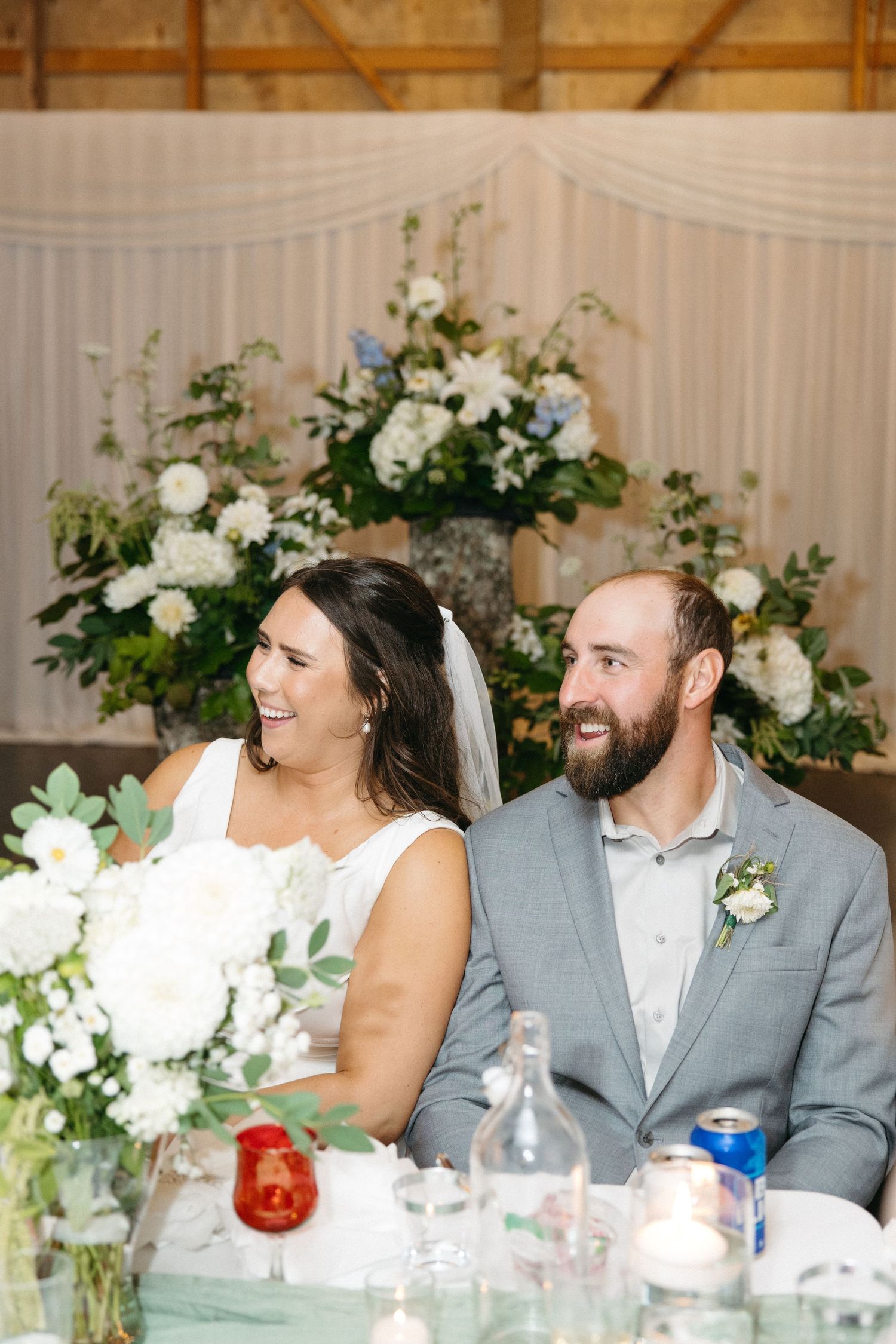Newlyweds laugh together at their reception table decorated with white floral arrangements.