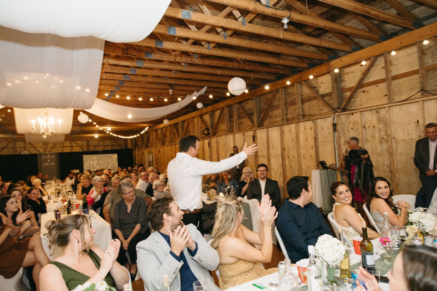 Wedding reception crowd cheers in rustic barn venue with string lights overhead.