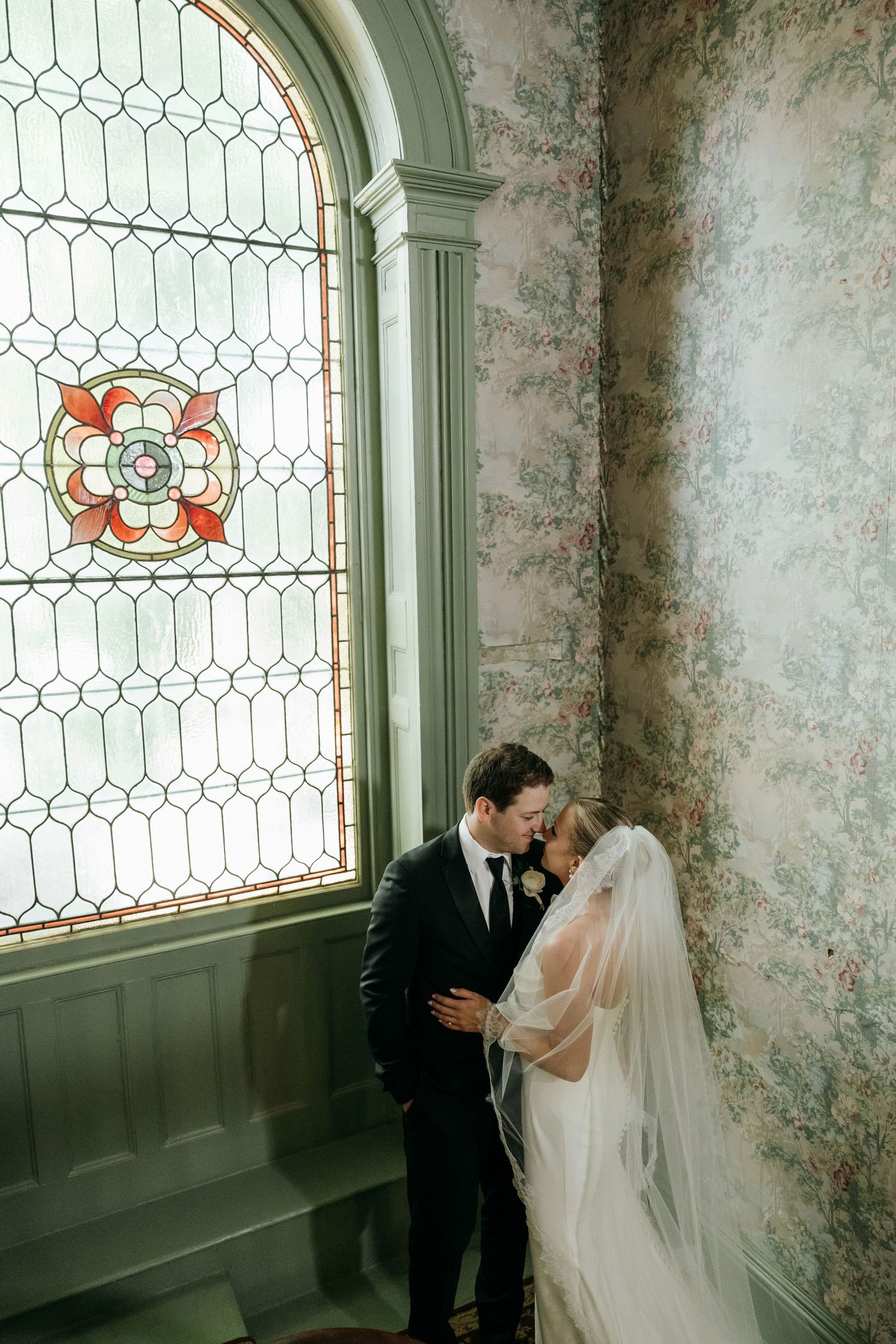 Romantic wedding moment by vintage stained glass window with floral pattern and textured wallpaper.