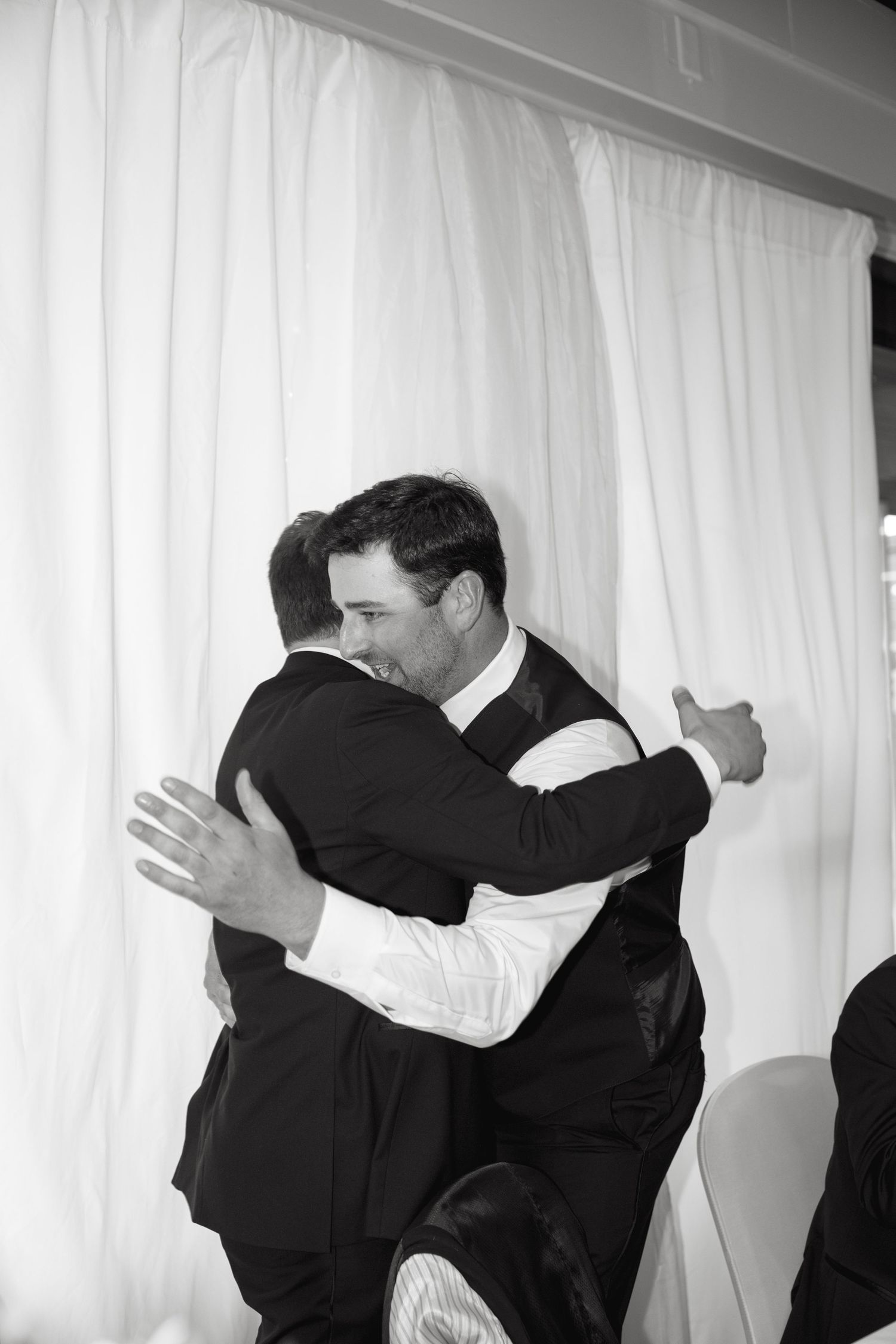 Two people share an emotional embrace against white curtains in black and white.