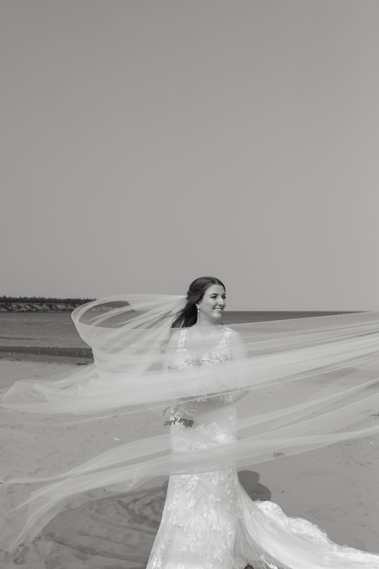 A flowing white wedding veil catches the ocean breeze on a sandy beach.