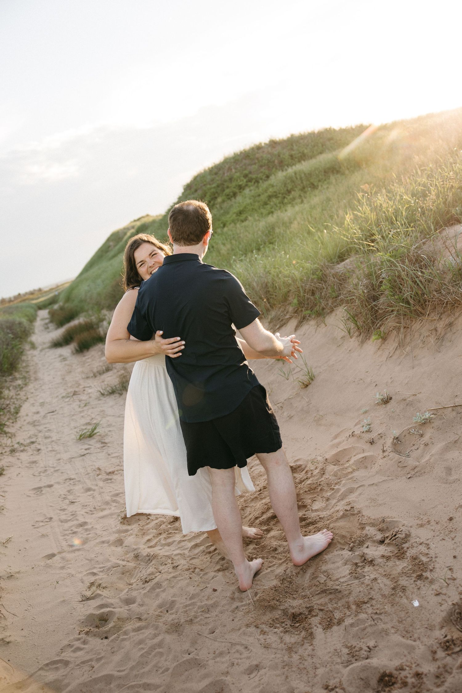 Couple embracing and dancing barefoot on sandy beach dunes at golden sunset hour.