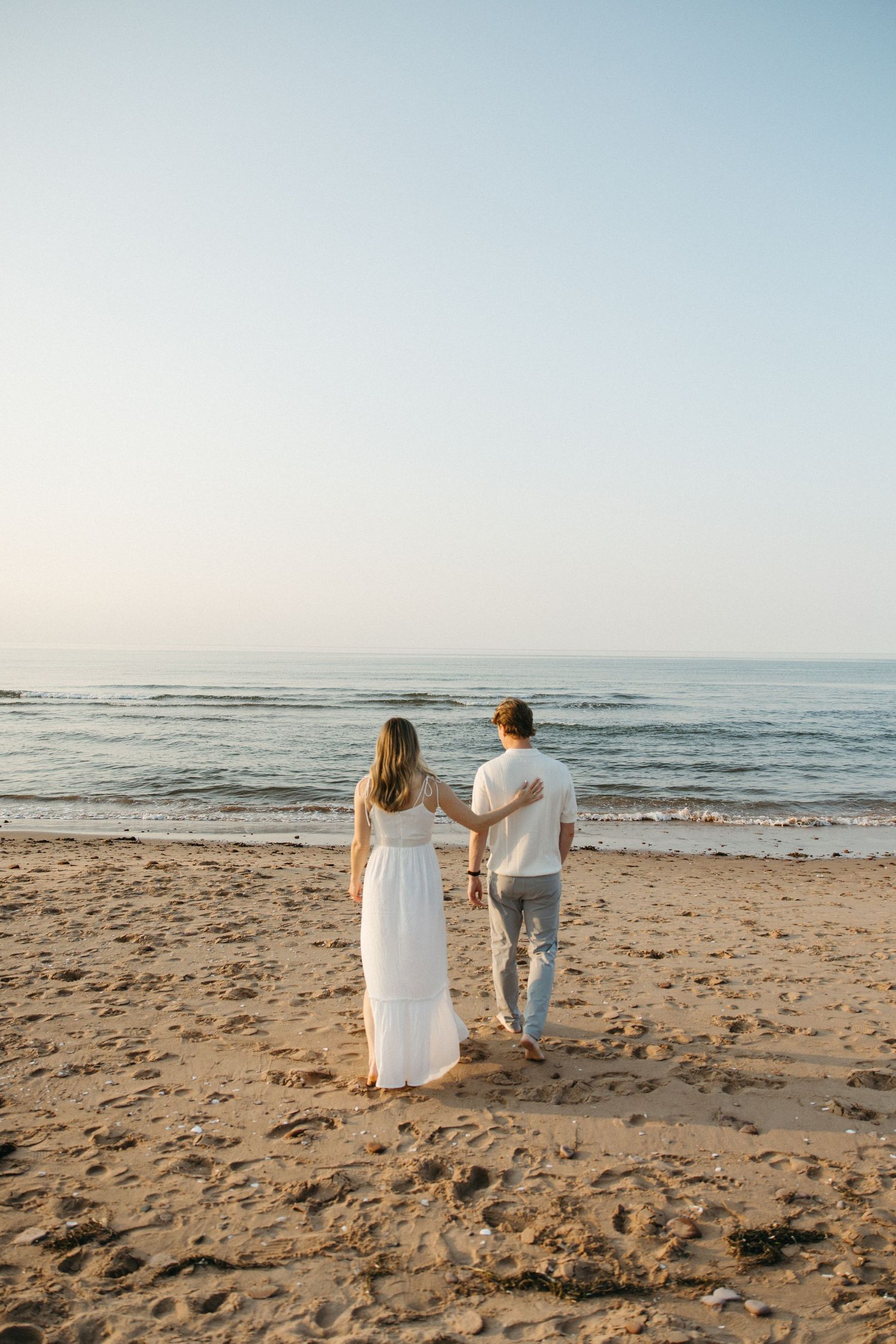 Couple in white outfits walking hand in hand toward calm ocean waves at sunset on sandy beach.