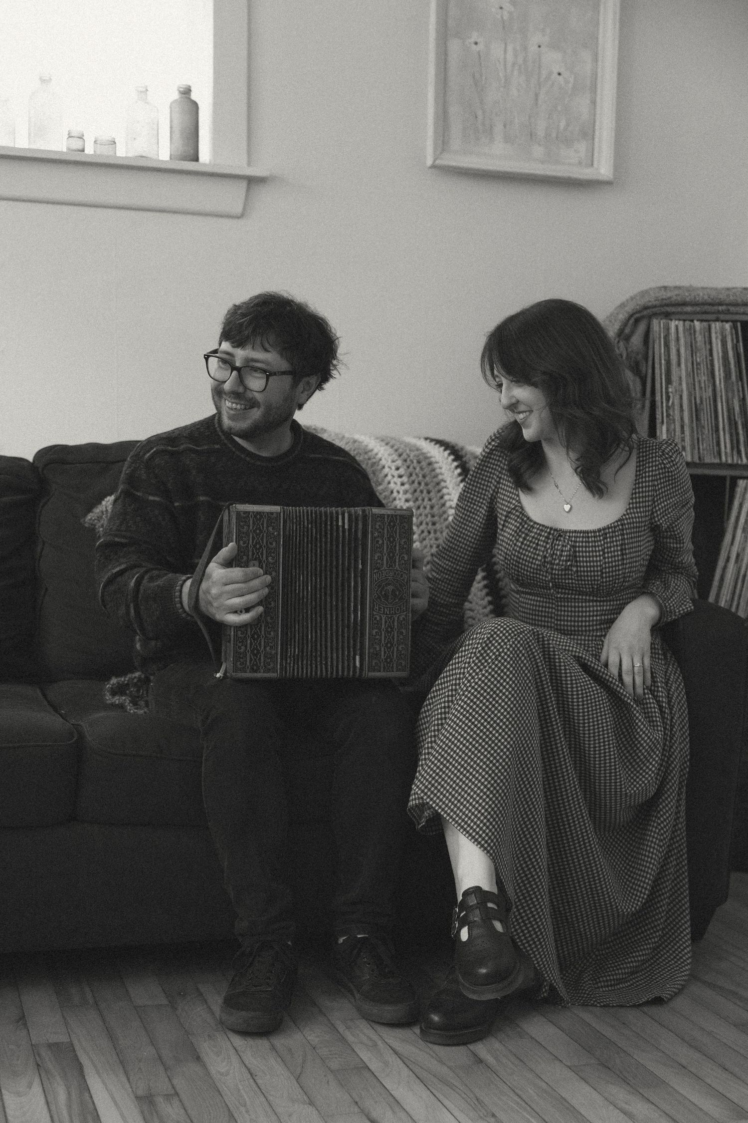A man playing accordion and a woman laughing together on a couch in a cozy living room, black and white photo.