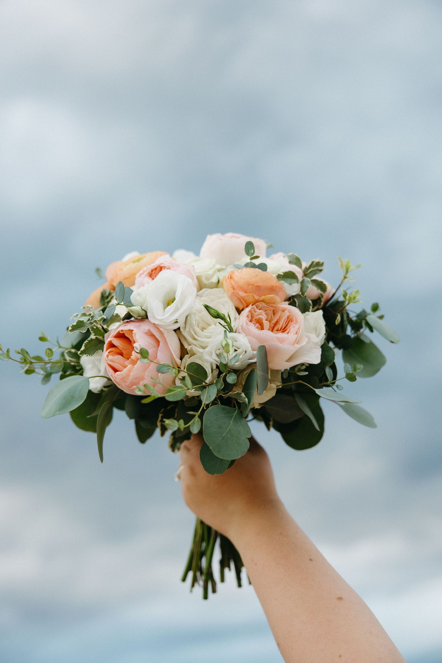 Hand holding a bridal bouquet of peach and white roses with eucalyptus against a cloudy sky.