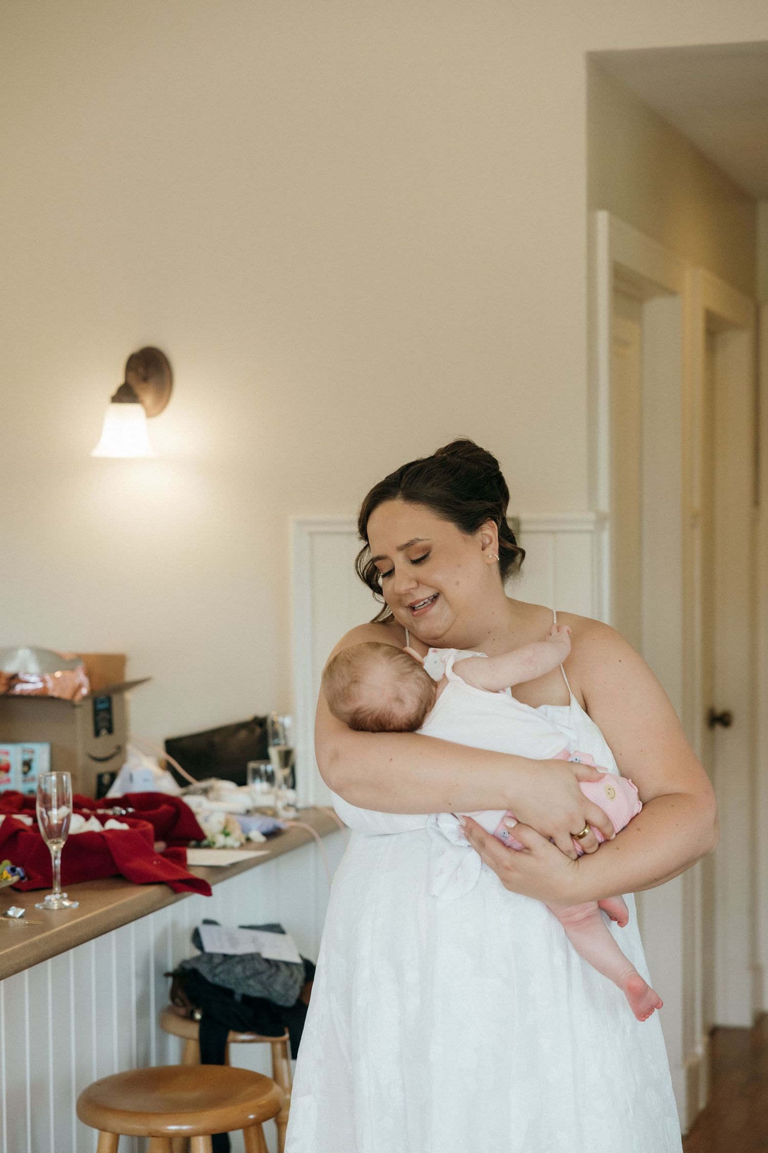 Bride in white wedding dress tenderly holding a newborn baby in a warmly lit getting-ready room.