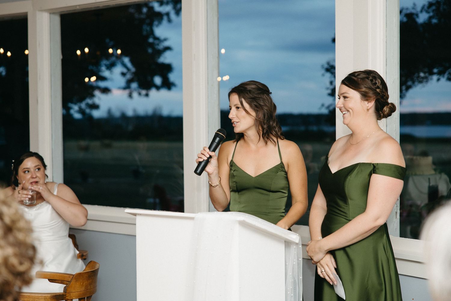 Two bridesmaids in green dresses giving a speech at a wedding reception podium near large windows.