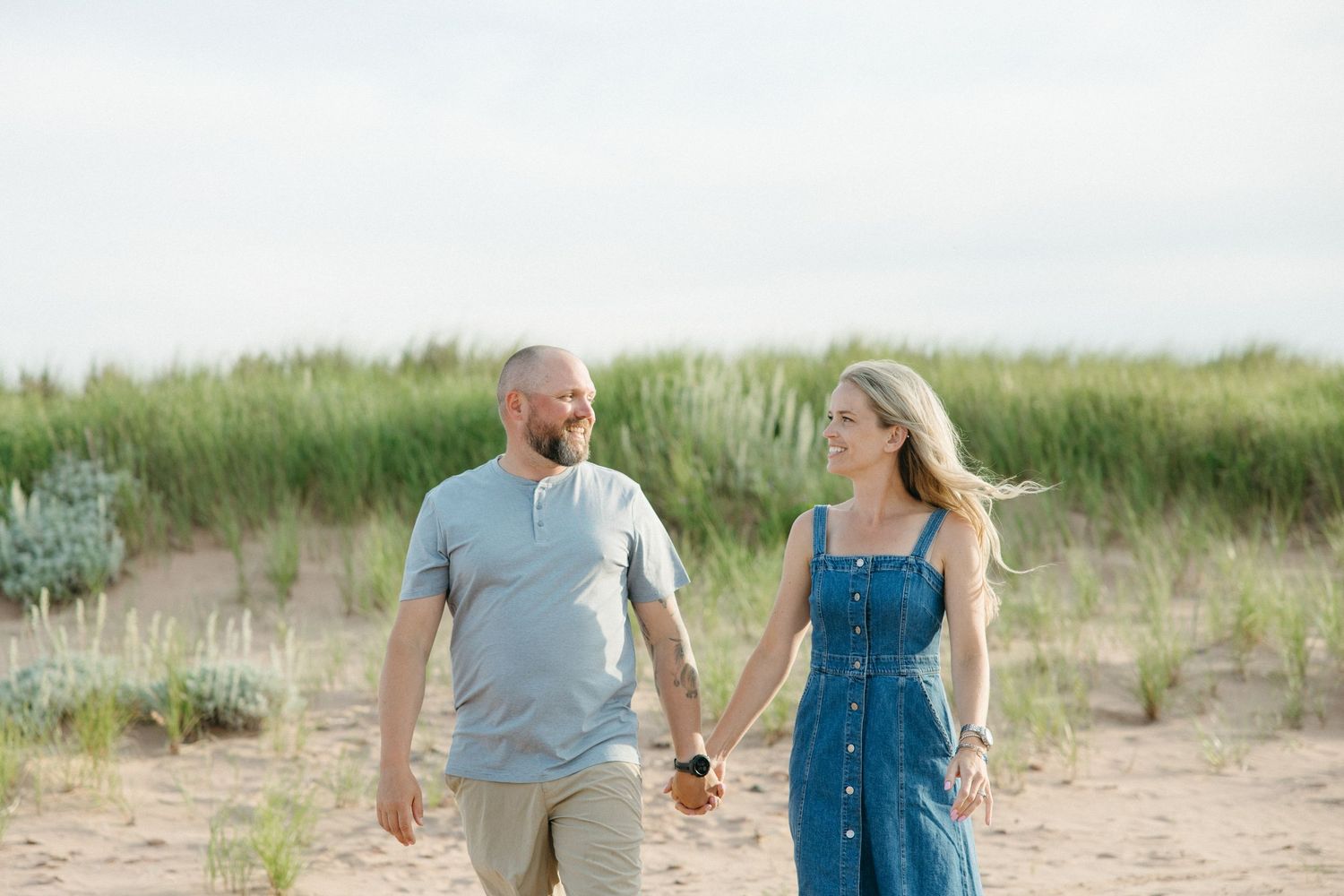Couple holds hands and smiles at each other while walking on sandy beach with green dunes behind them.