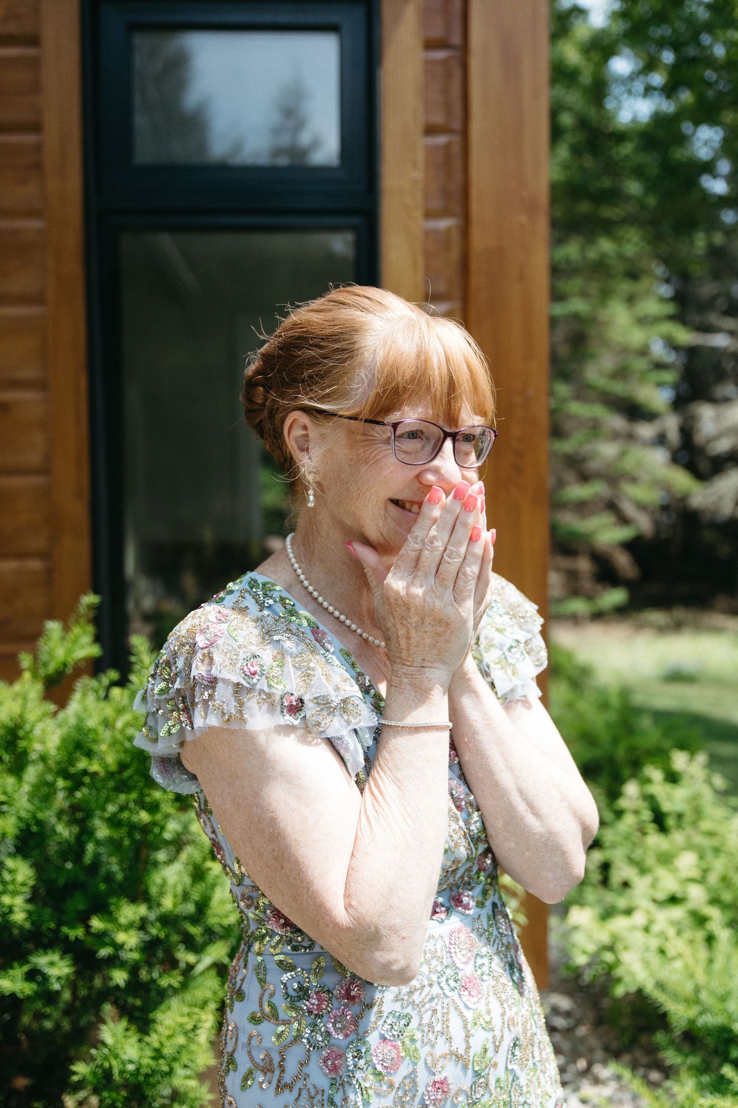 Elderly woman in floral dress and pearl necklace covering mouth with hands in surprise outdoors.