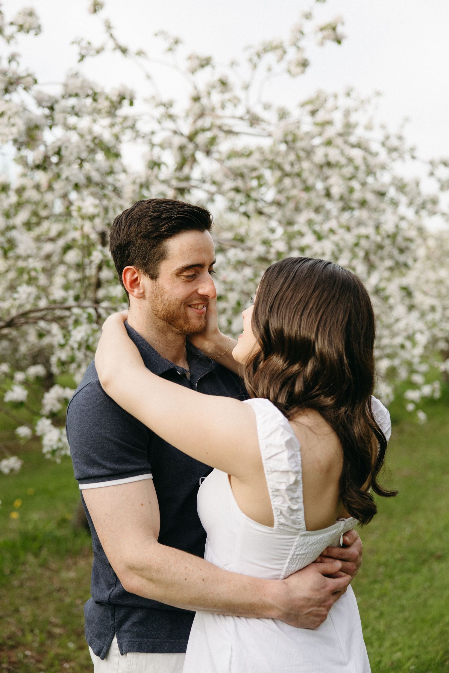 A smiling man holds a woman in a white dress while standing before a flowering tree.