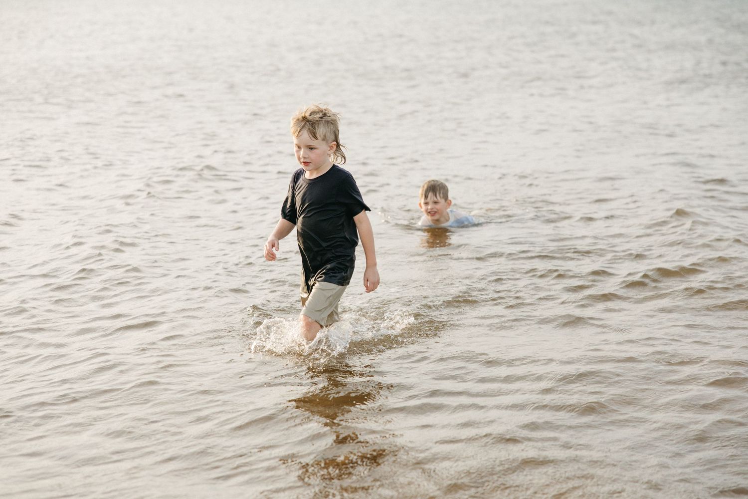 Two boys playing in shallow lake water, one wading and one swimming on a sunny day.