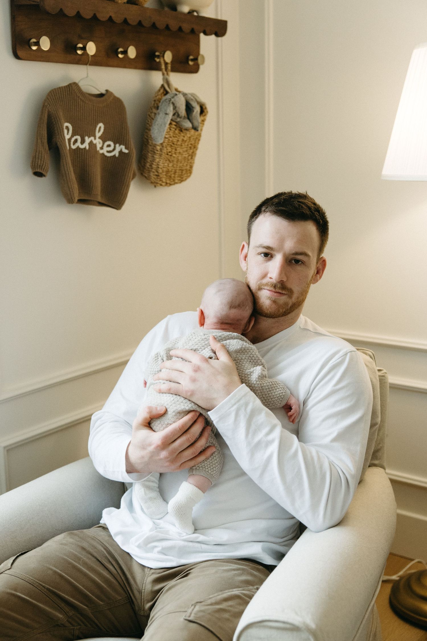 A tender moment in a nursery with a brown sweater labeled 'Parker' hanging on wall hooks above.