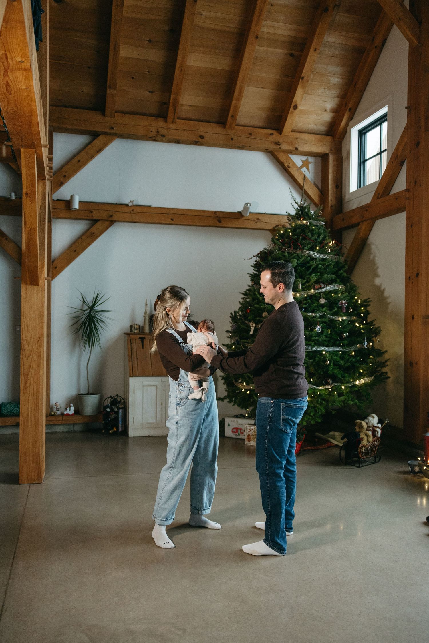 A couple stands together in a rustic room with exposed wooden beams and Christmas trees.