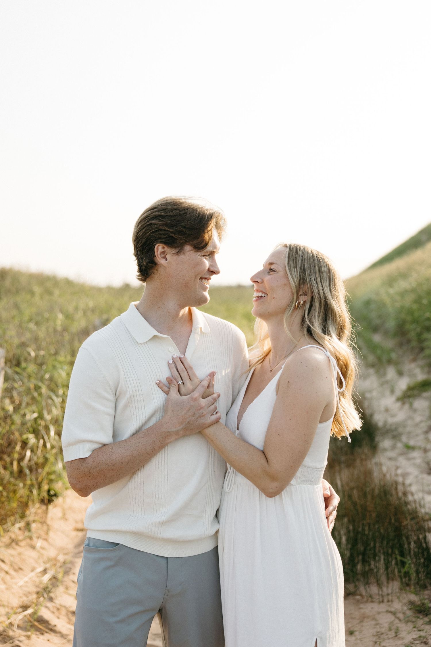 Couple smiling at each other on sandy beach dunes, woman in white dress holding man's hands at golden hour.