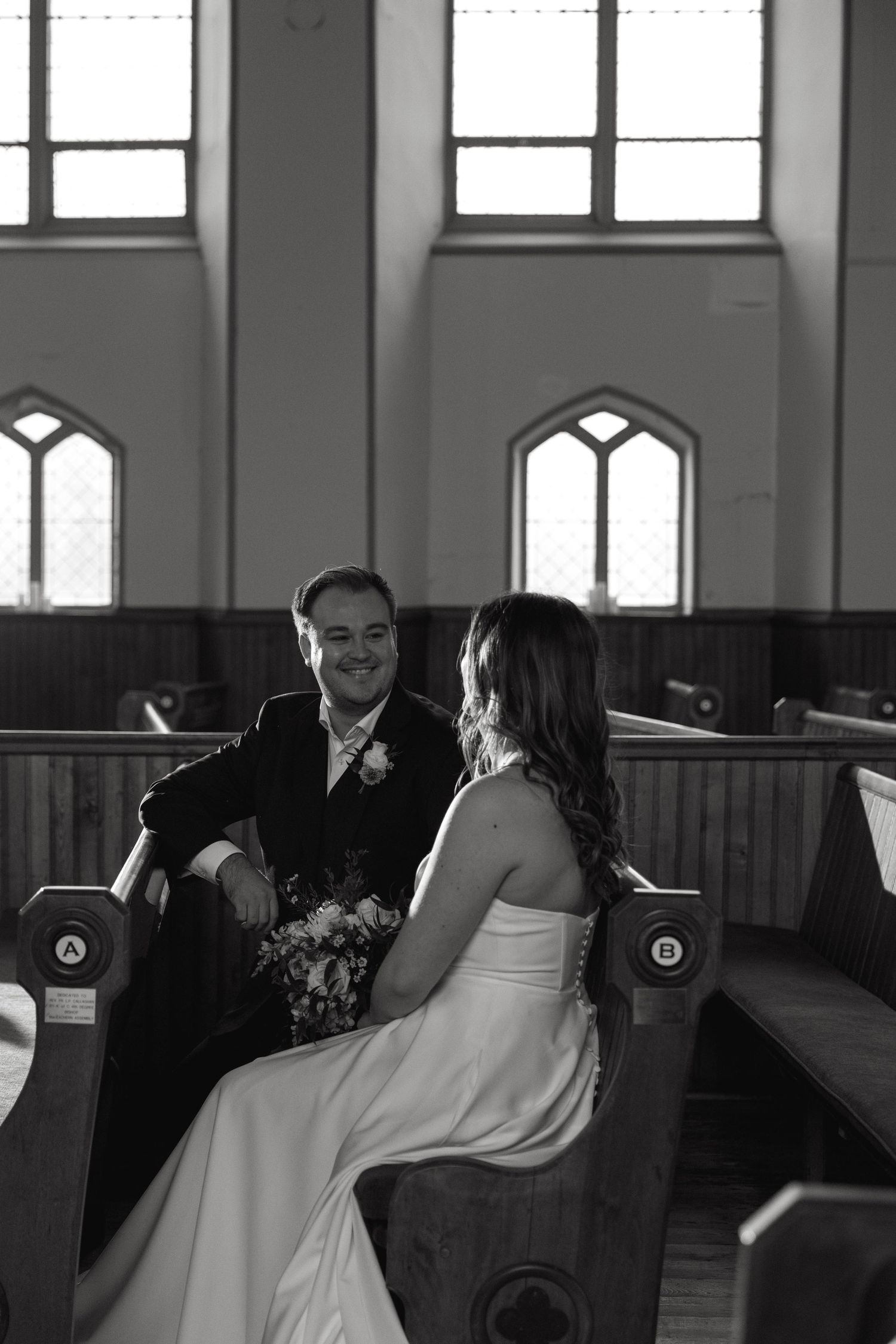 A black and white photo of a couple seated in wooden church pews with Gothic windows in the background.