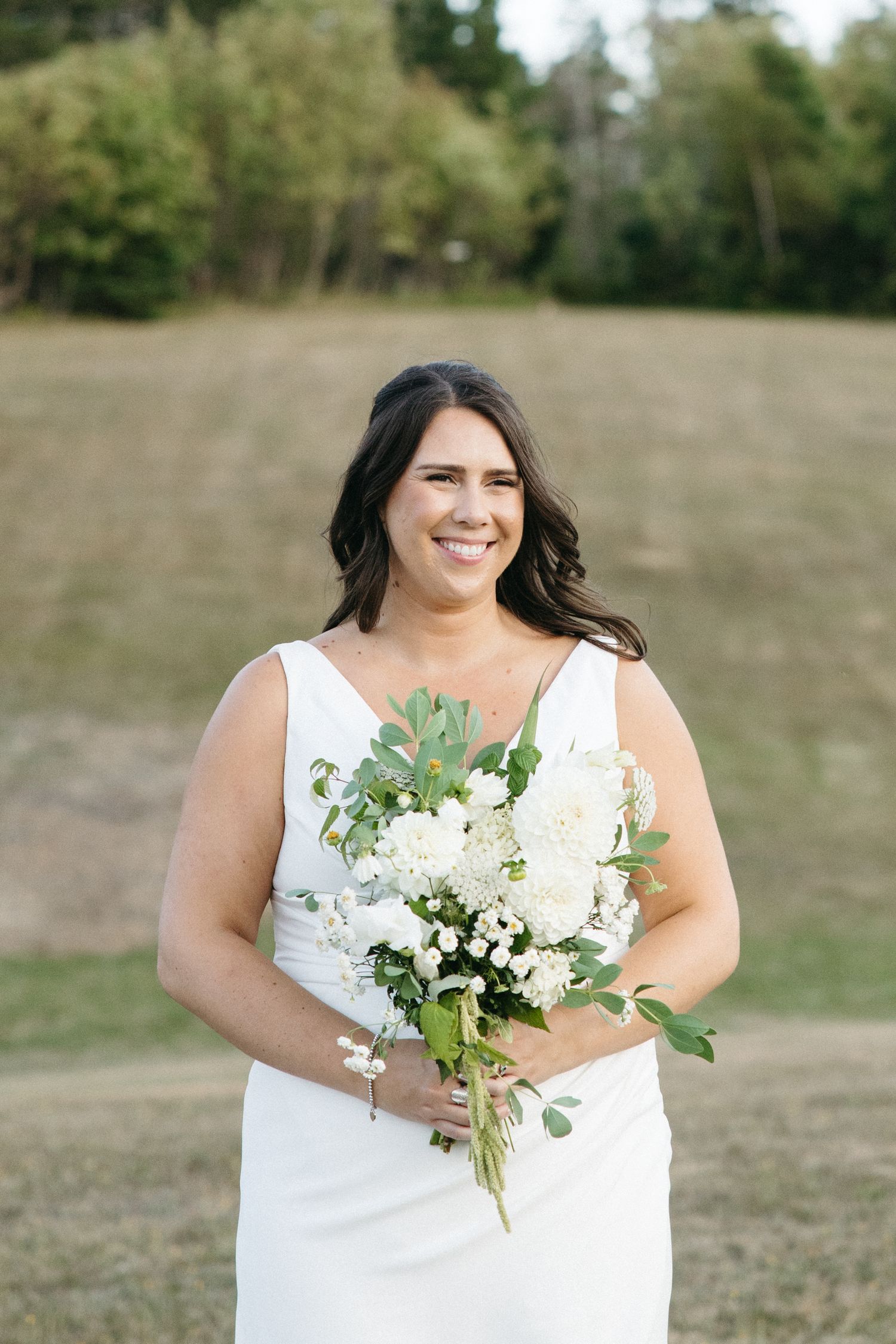 Bride in white dress holding elegant white and green floral bouquet outdoors.