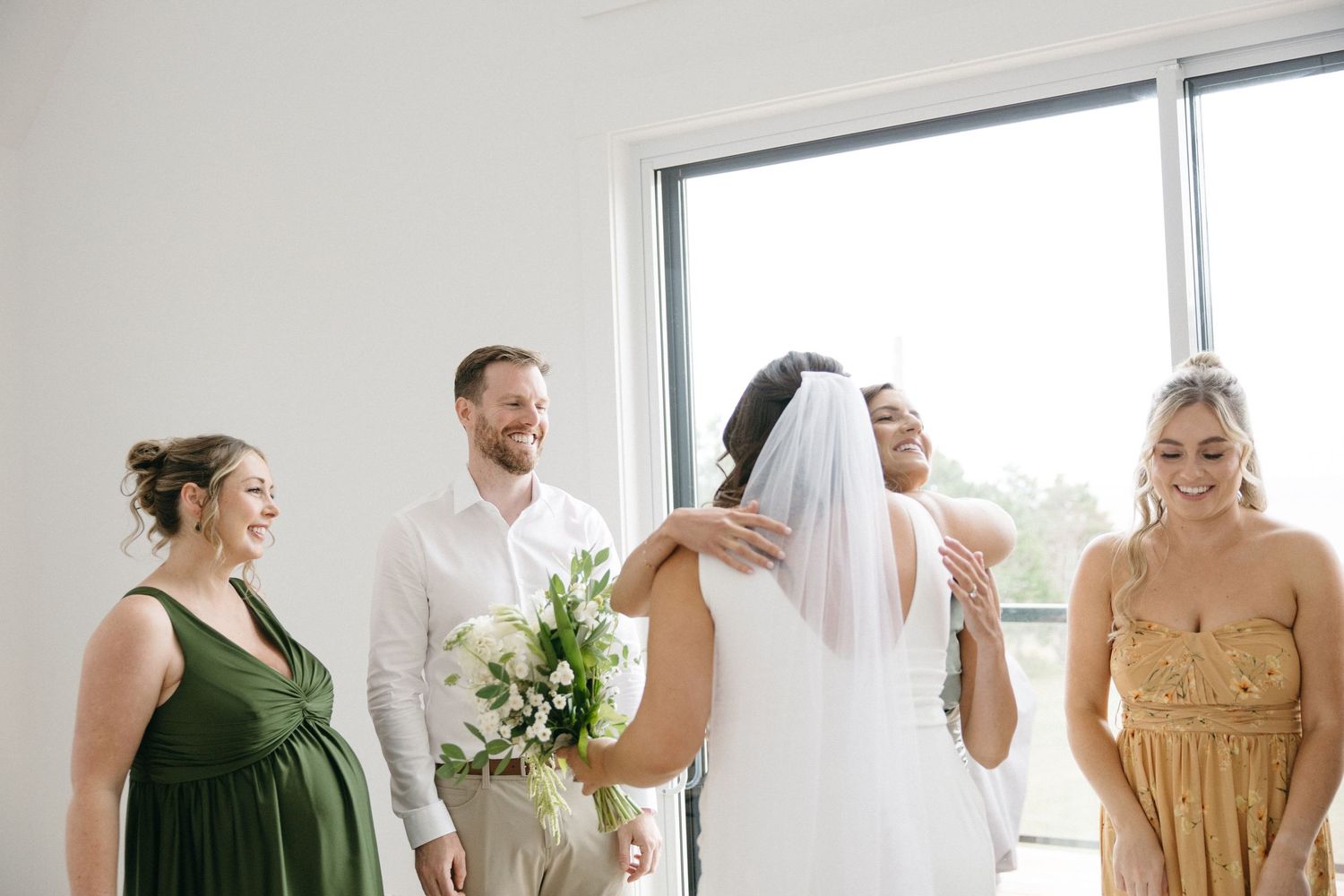 Wedding party members in earth-toned dresses gather in a minimalist white room.