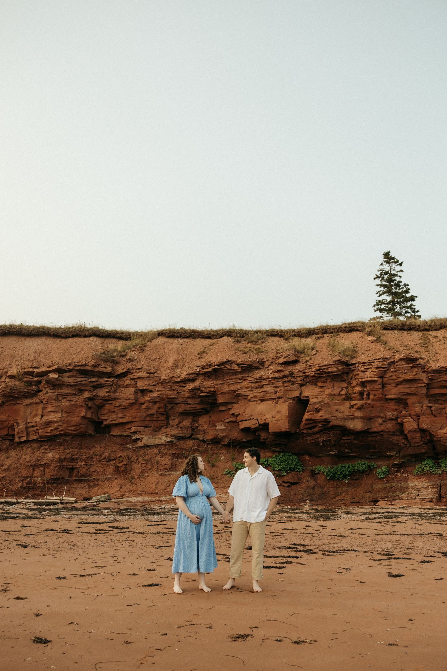 Two people walk along a rocky shoreline with dramatic red sandstone cliffs and pine trees.