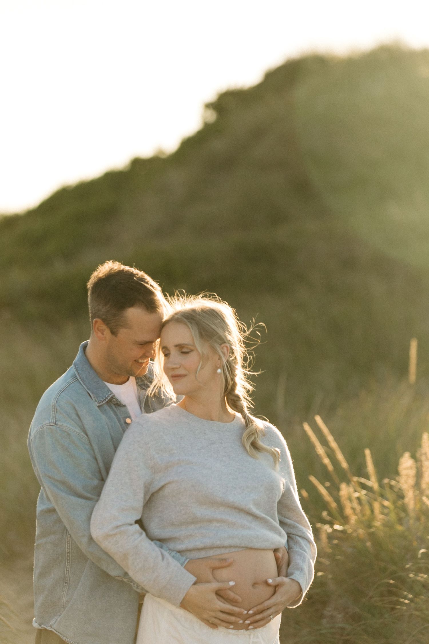 Intimate maternity photo of a couple embracing in golden sunlight on a hillside.