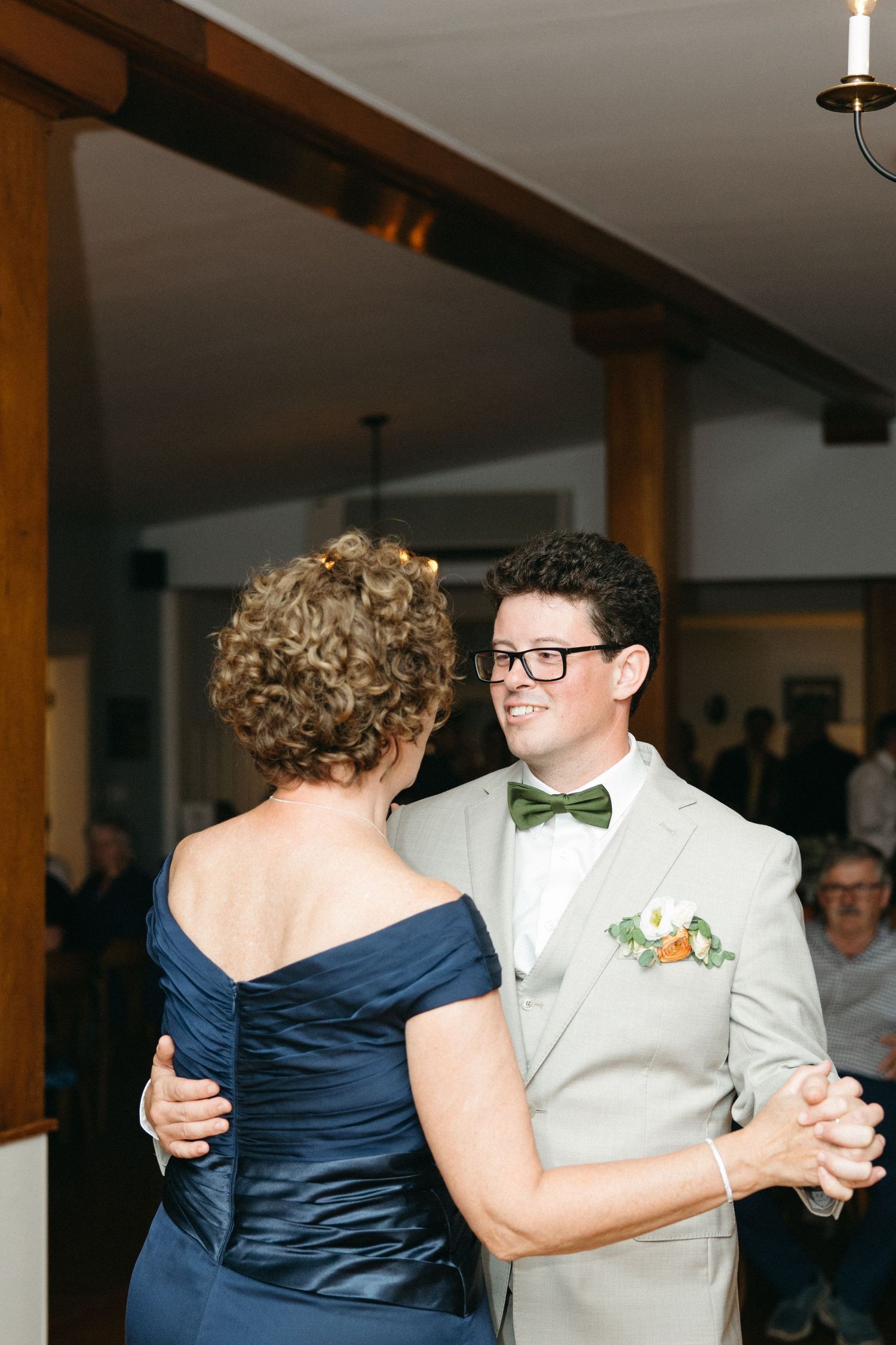 Groom in gray suit and green bow tie shares mother-son dance at elegant wedding reception.