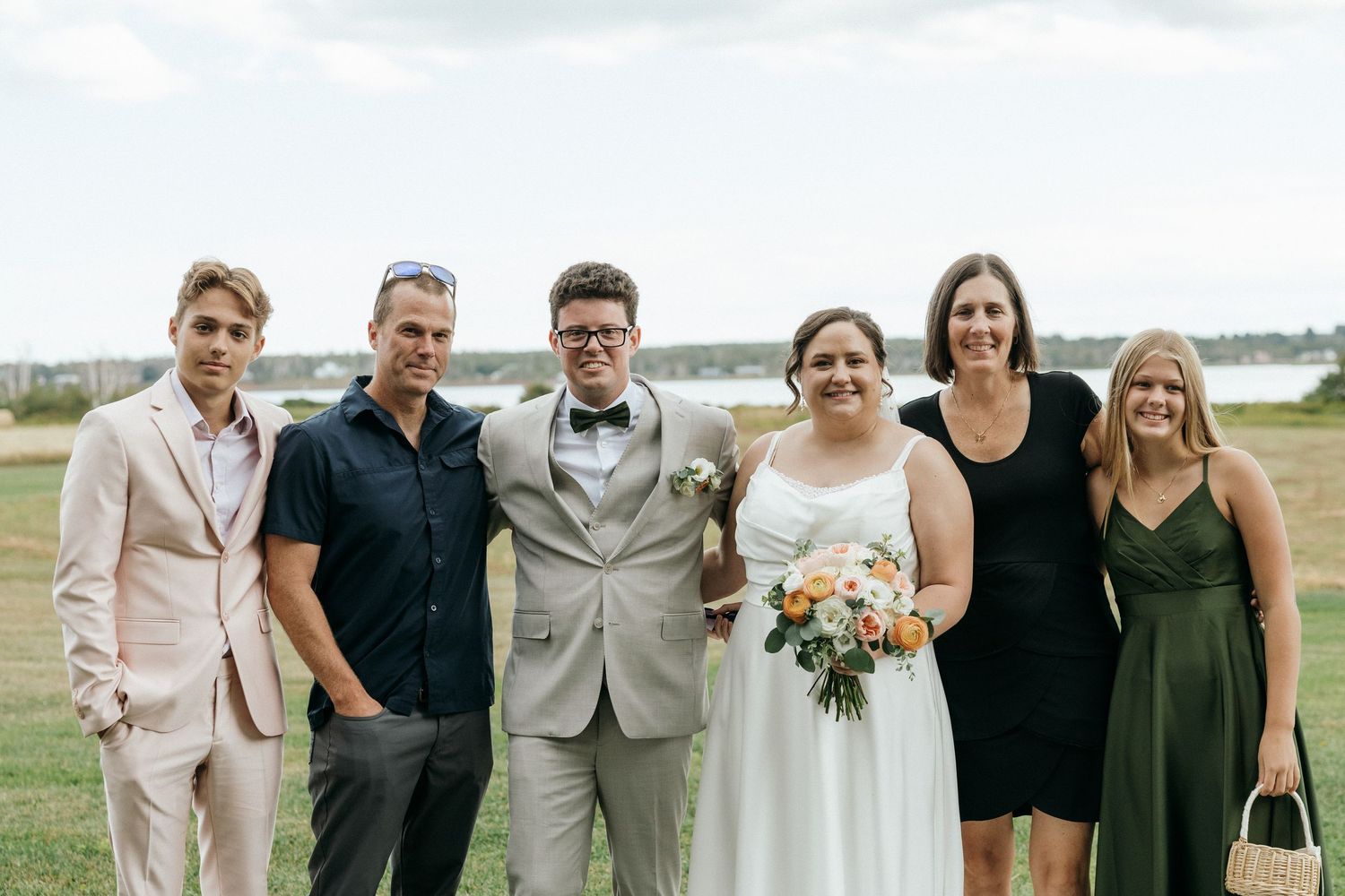 Bride and groom pose with four wedding guests on grassy lawn with waterfront view in background.