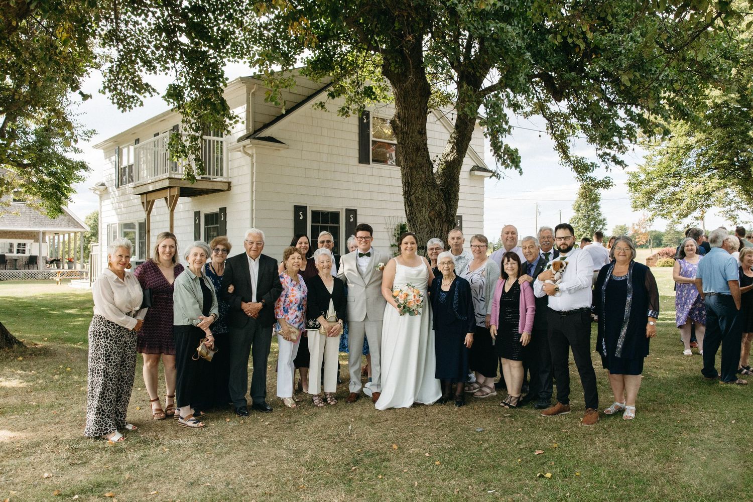 Wedding party and guests pose for group photo under large tree outside white venue.