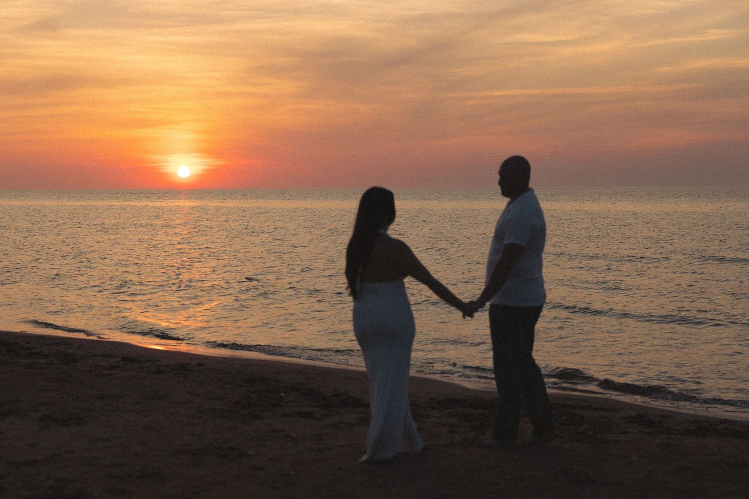 Romantic couple holding hands on a beach at sunset with vibrant orange sky reflecting on calm water.