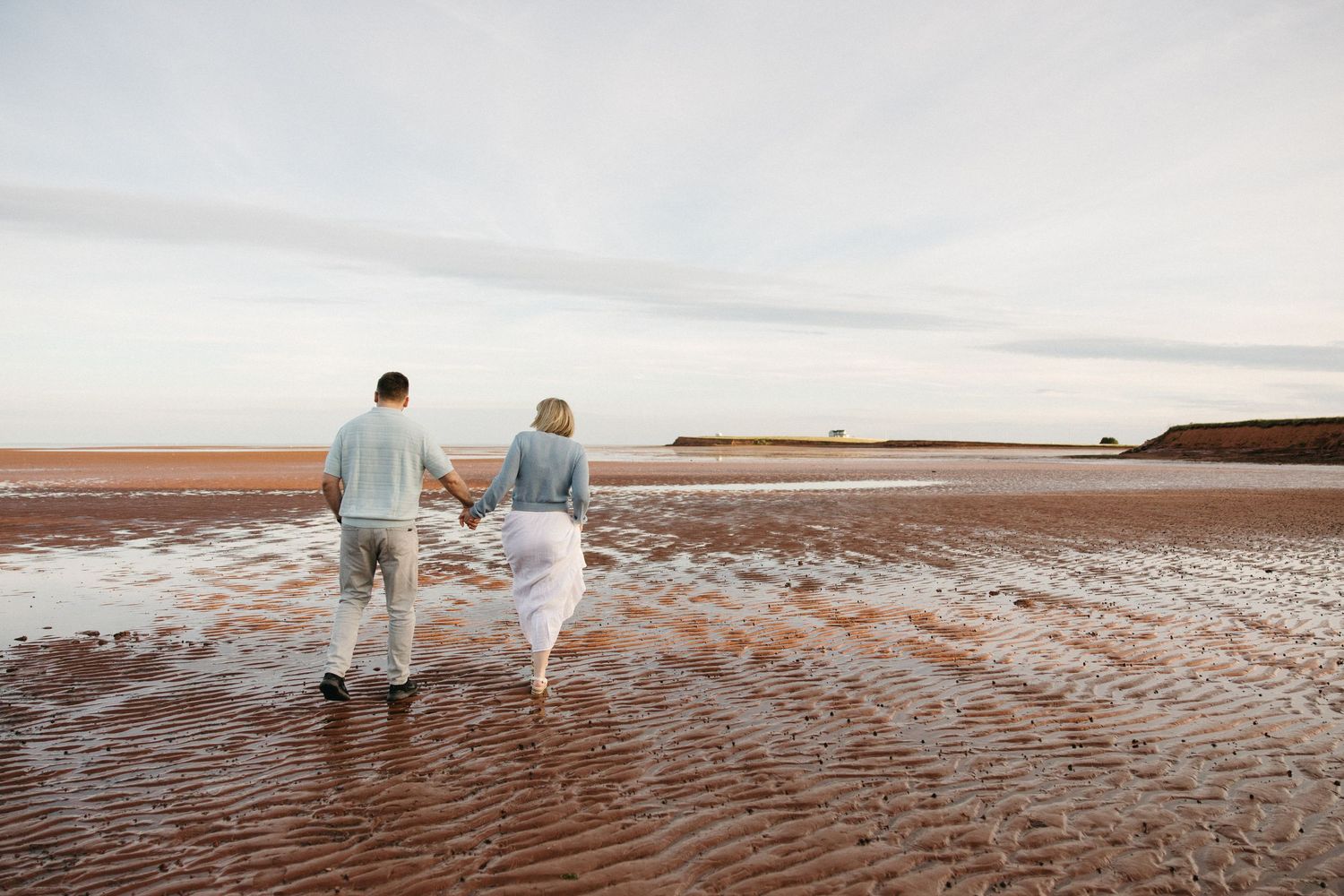 Couple holding hands walking on a red sand beach at sunset, viewed from behind, wearing casual white and grey outfits.
