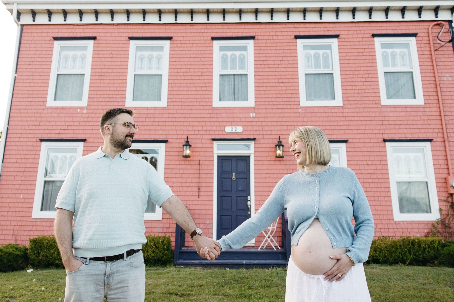 Pregnant couple holding hands in front of a red brick house, smiling at each other during a maternity photo shoot.